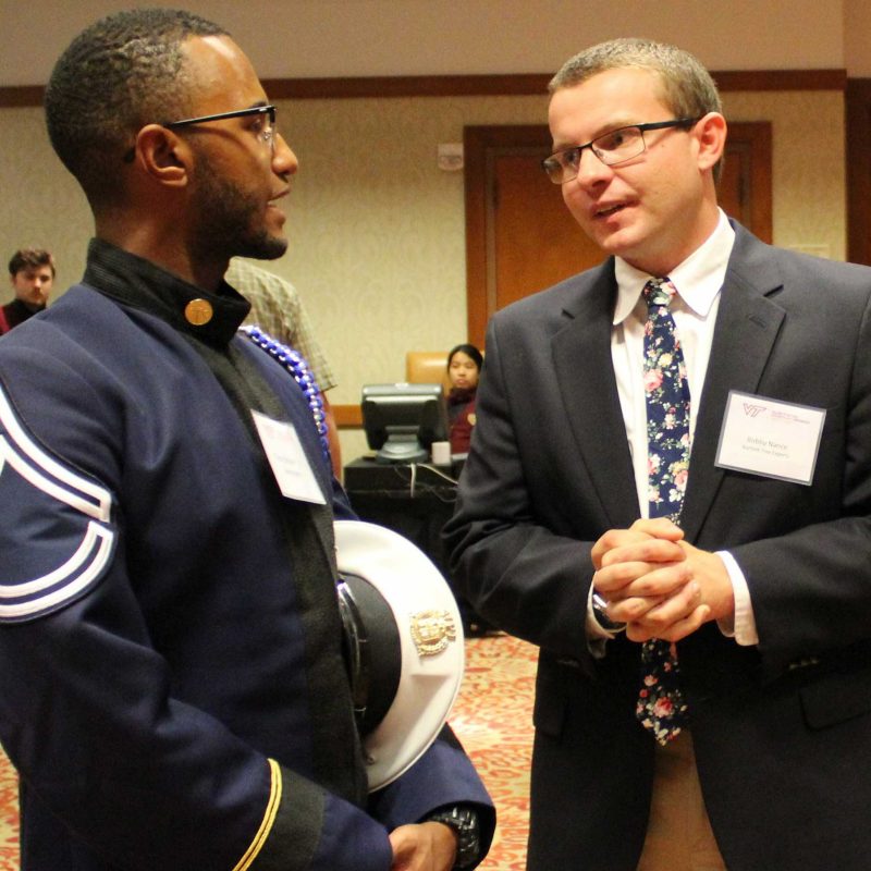 A young man wearing a military uniform talks with another young man wearing a tie and sports jacket. Several people are visible in the background.