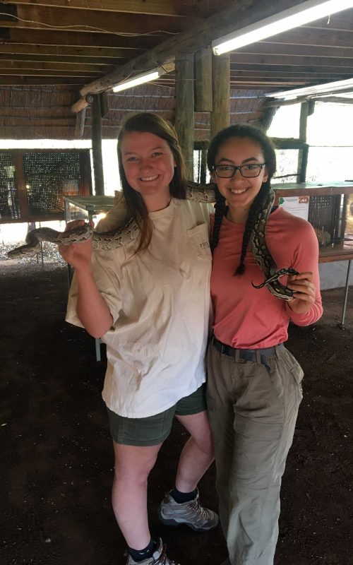 Two young women stand side-by-side, jointly holding a large snake over their shoulders.