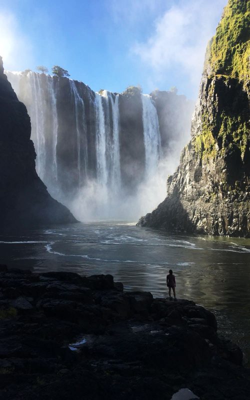 A person stands near the base of a large waterfall.