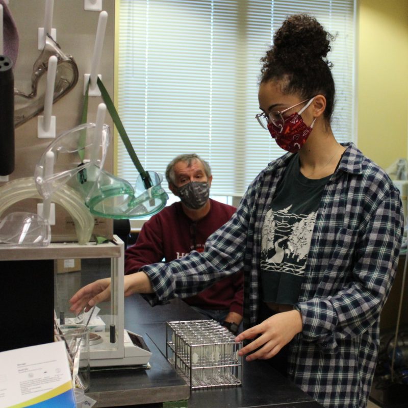 A young woman wearing a face mask in a lab holding a bank of tubes with a professor wearing a face covering in the background.