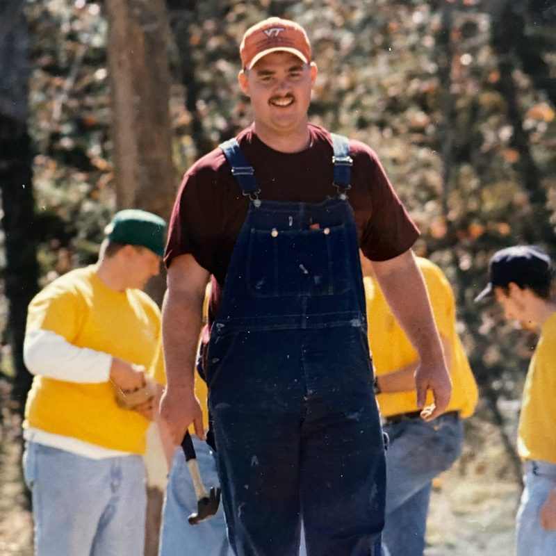 A young man smiling, wearing overalls, t-shirt, and hat, holding a hammer by his side.