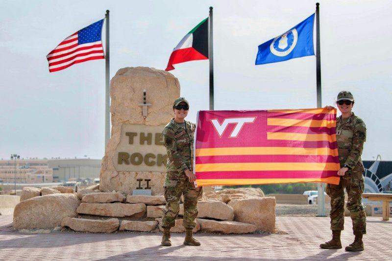 Two U.S. soldiers holding a VT orange and maroon stripped flag in front of The Rock II military base entrance, a sword in stone.