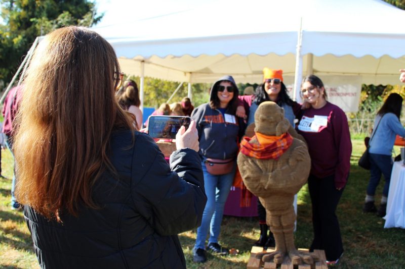 Three female alumnae pose behind a HokieBird constructed of corrugated paperboard while another female takes their photo with a mobile device.