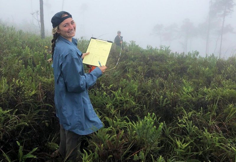 A graduate student smiling in a field holding a measuring tape and clipboard.
