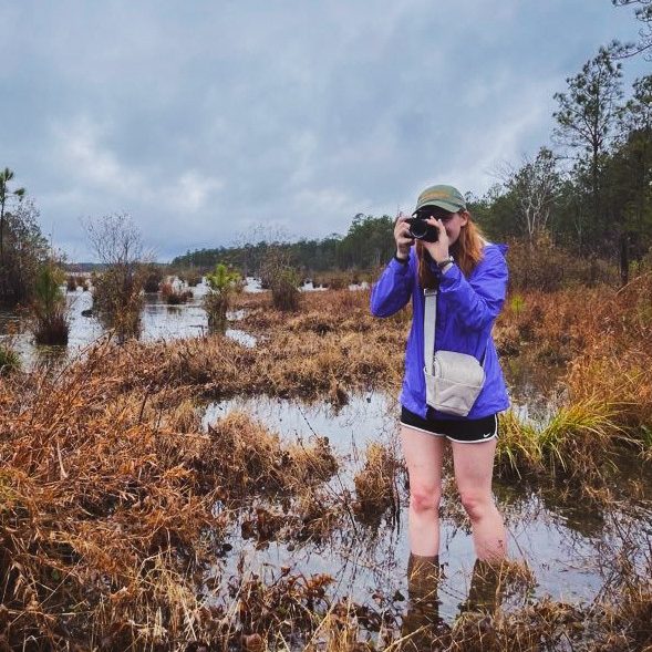 Sydney Haney standing in a bog with a camera up to her face.