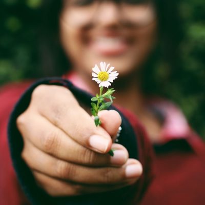 A young woman out of focus holding a flower toward the camera in focus.