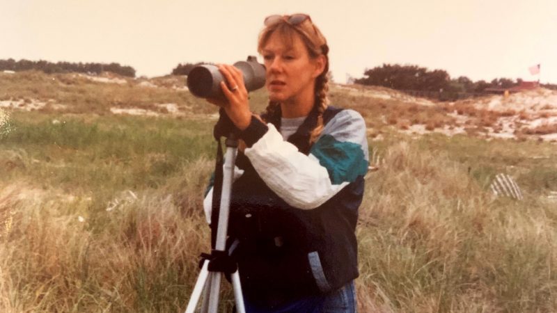 A woman on a barrier island looking through the lens of a spotting scope on a tripod.
