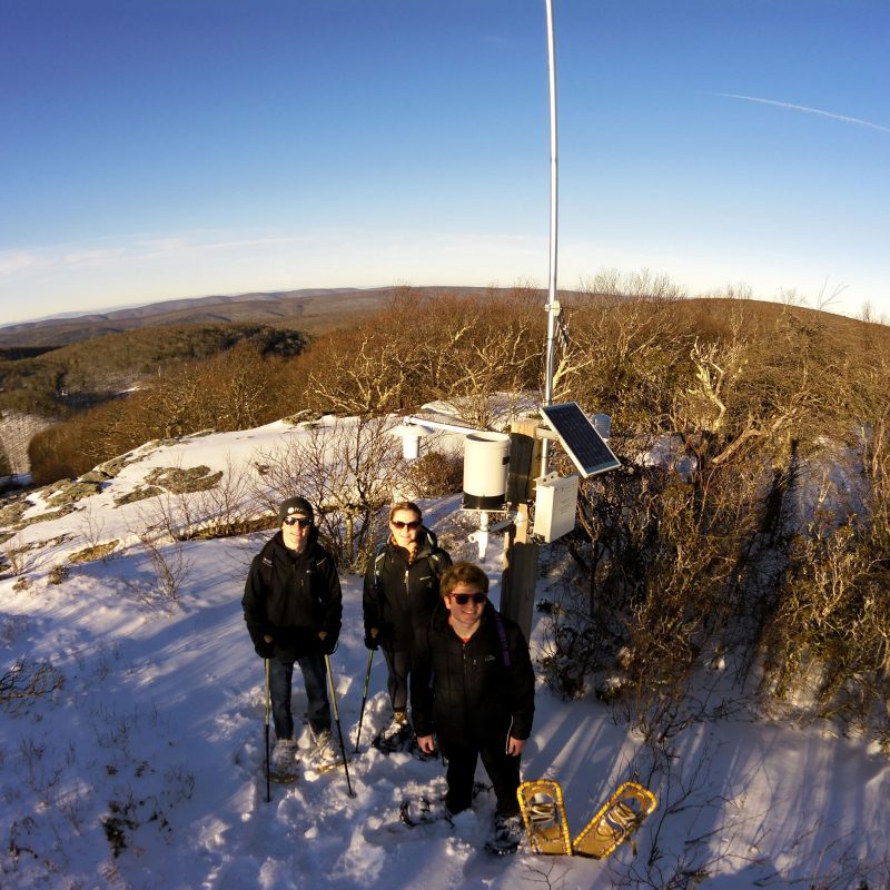 Three people stand in front of a weather station on a mountaintop, with snow on the ground.
