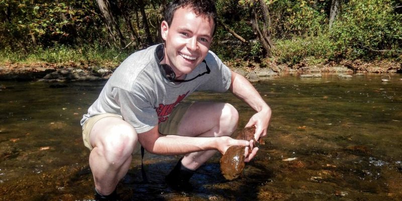 Brian Case holds a large salamander while crouching in a forest stream