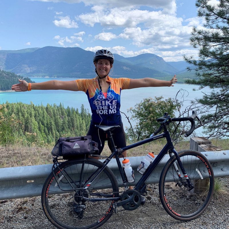 A female transcontinental bicyclist for Bike the U.S. for M.S. with outstretched arms a lake and tree covered mountains in the background.