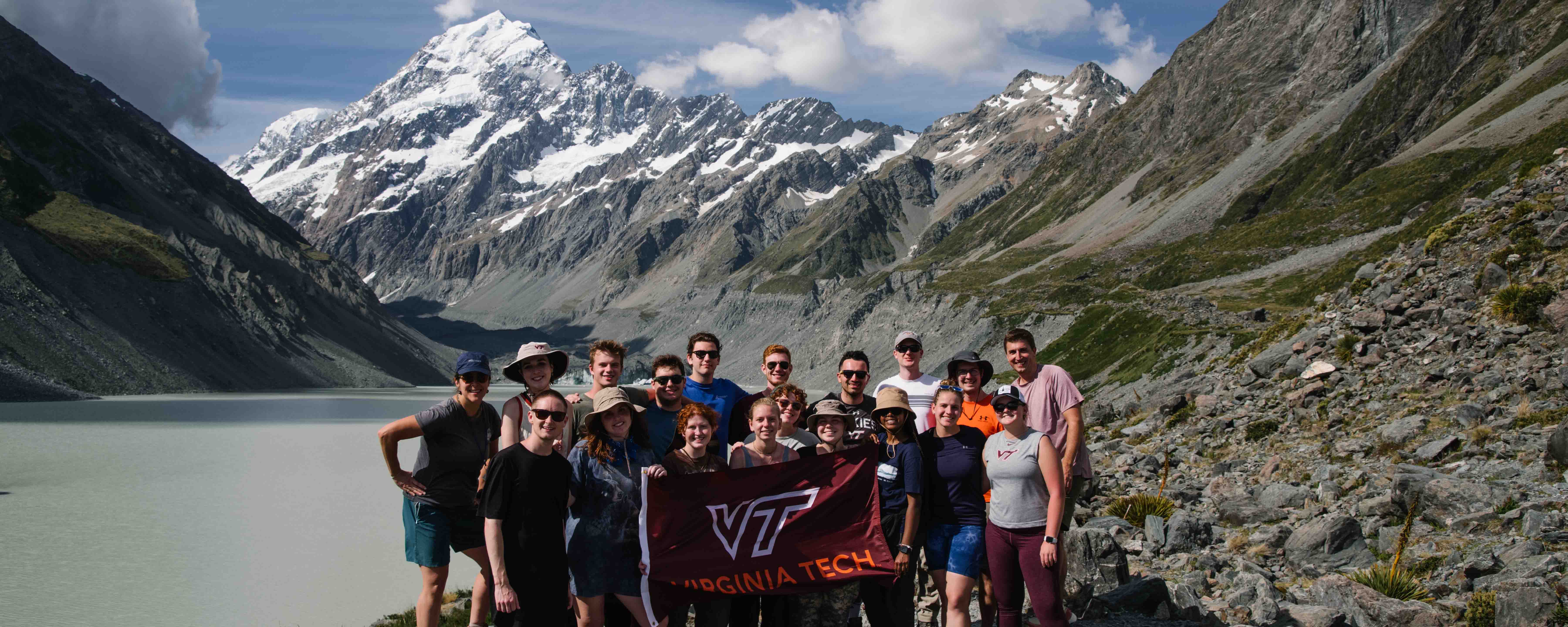 Students holding a Virginia Tech flag in front of a milky glacier lake and snow covered, skyscraping mountain peak.