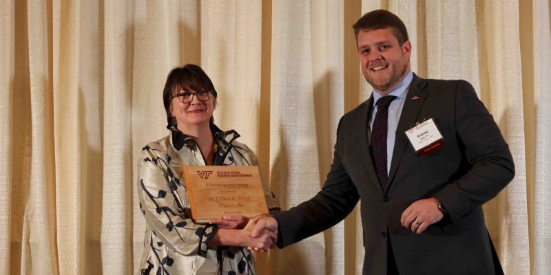 Bettina Ring holds an engraved wooden plaque while shaking Andrew Ickes' hand. The engraving reads, "2024 Distinguished Alumna Presented to Bettina K Ring Class of 1986, March 27, 2024."