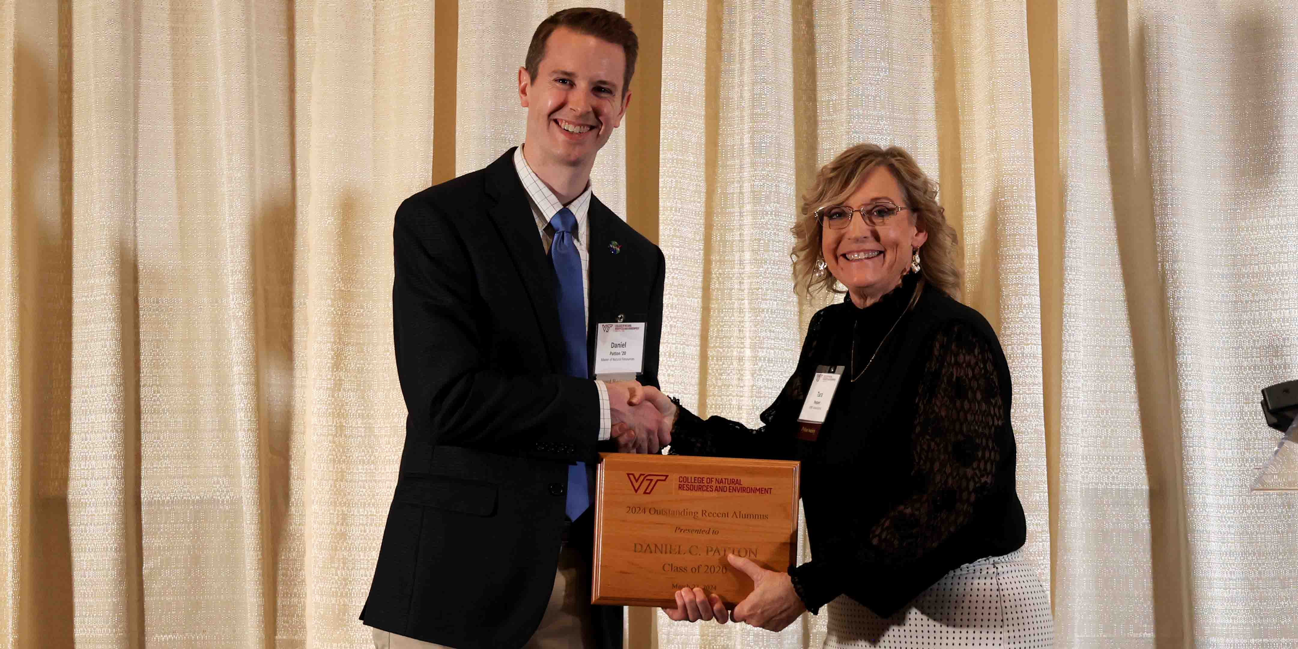 Daniel Patton holds an engraved wooden plaque while shaking Tara Nepper's hand. The engraving reads, "2024 Outstanding Recent Alumnus Presented to Daniel C Patton Class of 2020, March 27, 2024."