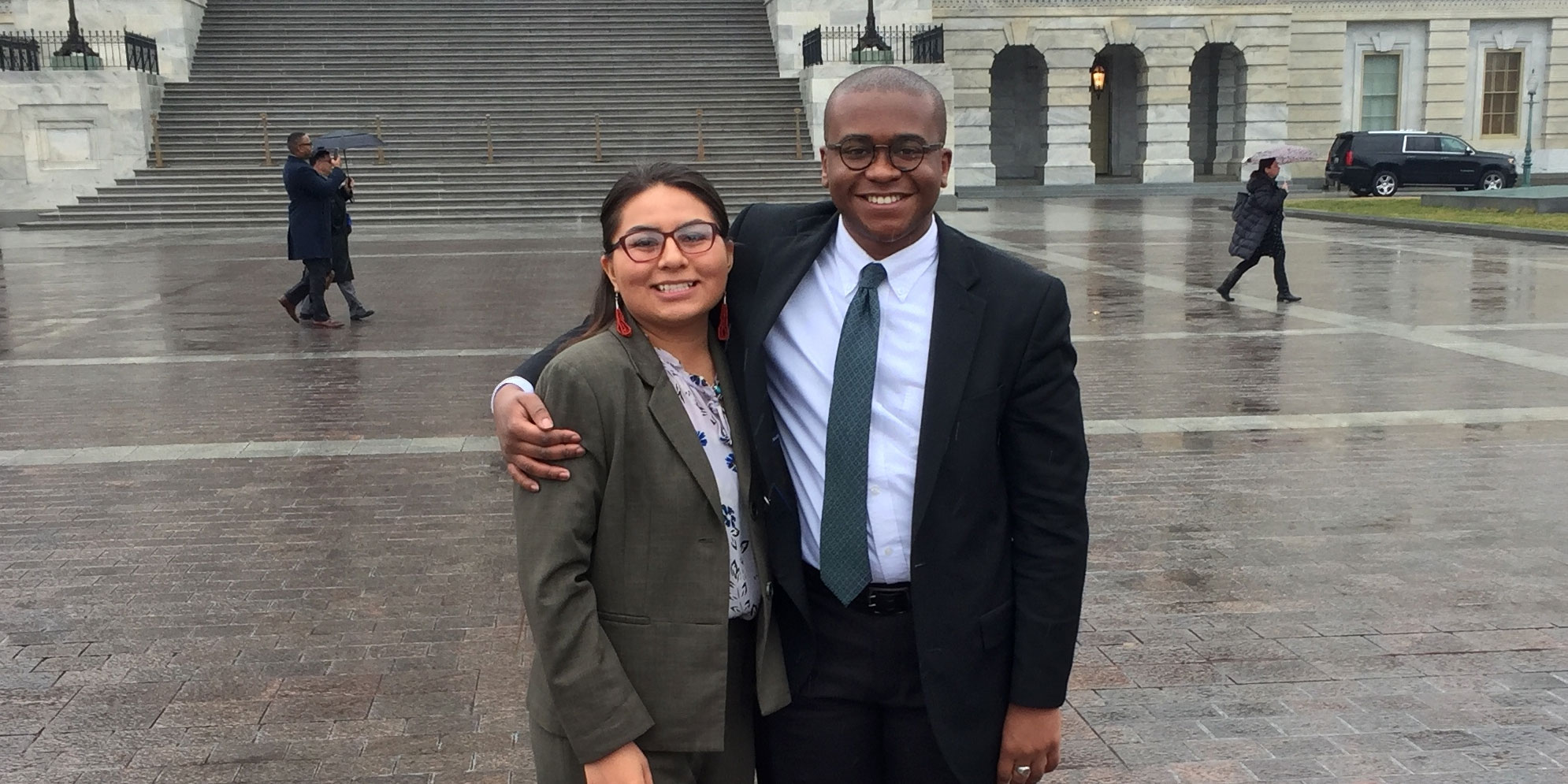 Two interns smiling and posing in front of the U.S Capitol Building on a rainy day.
