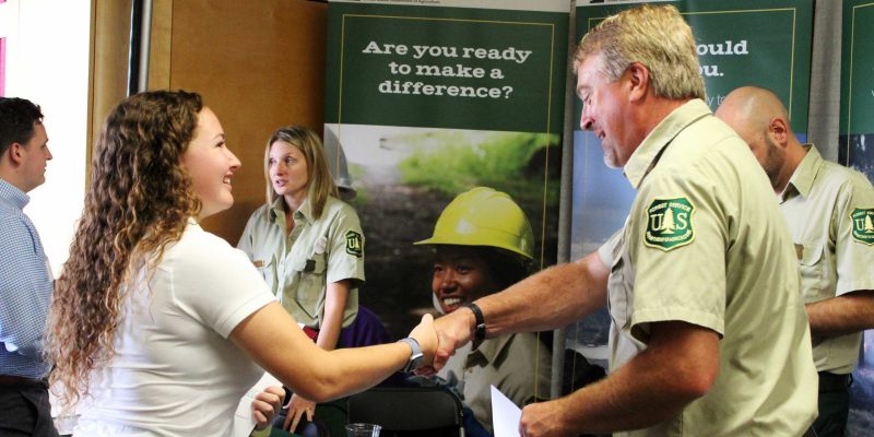 A young woman shakes hands across a table with a man wearing a green U.S. Forest Service uniform. Several other people are visible in the background.