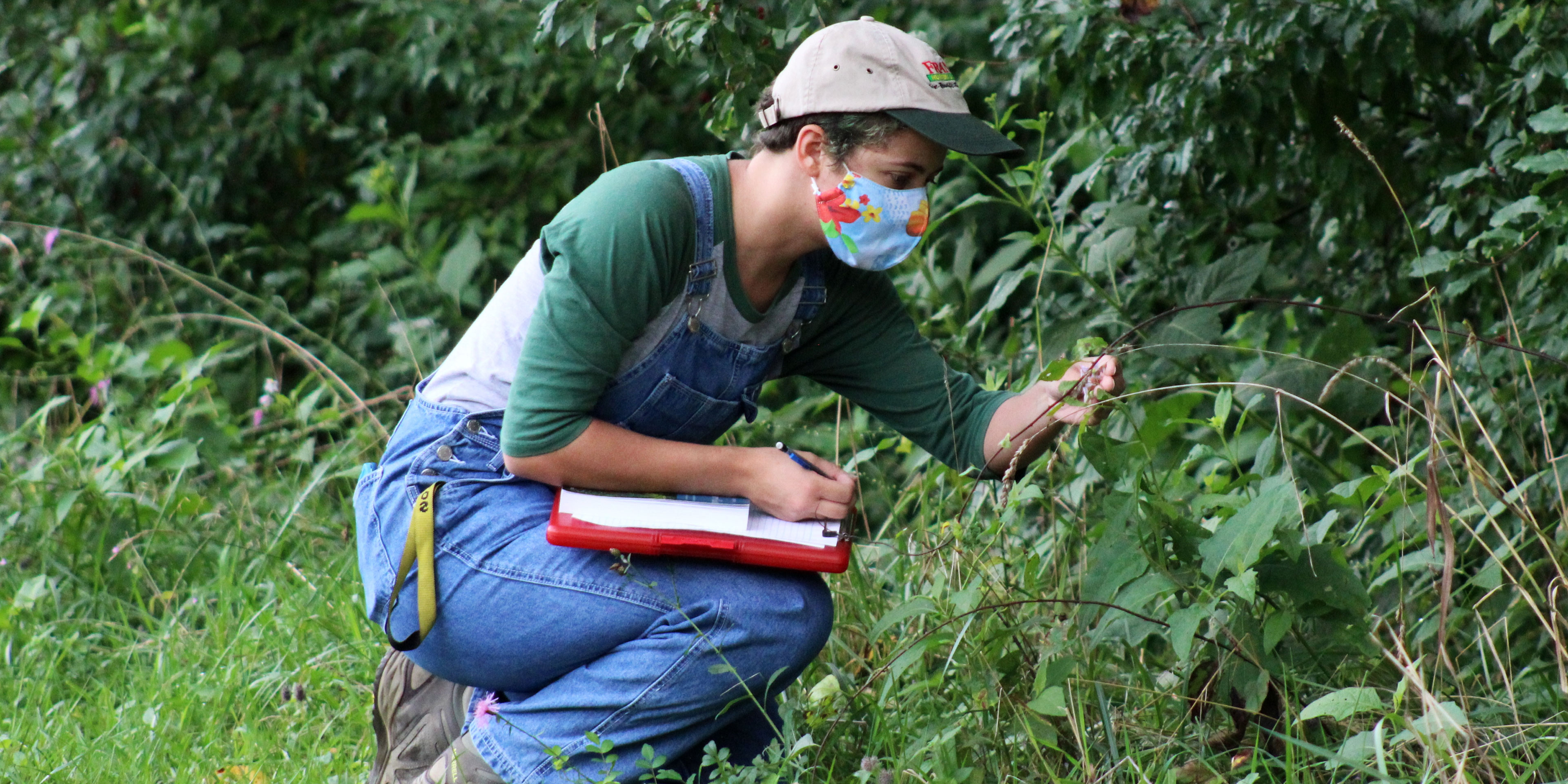 A young woman wearing a face mask outside crouches down and examines a plant with one hand while holding a pen and clipboard in the other.