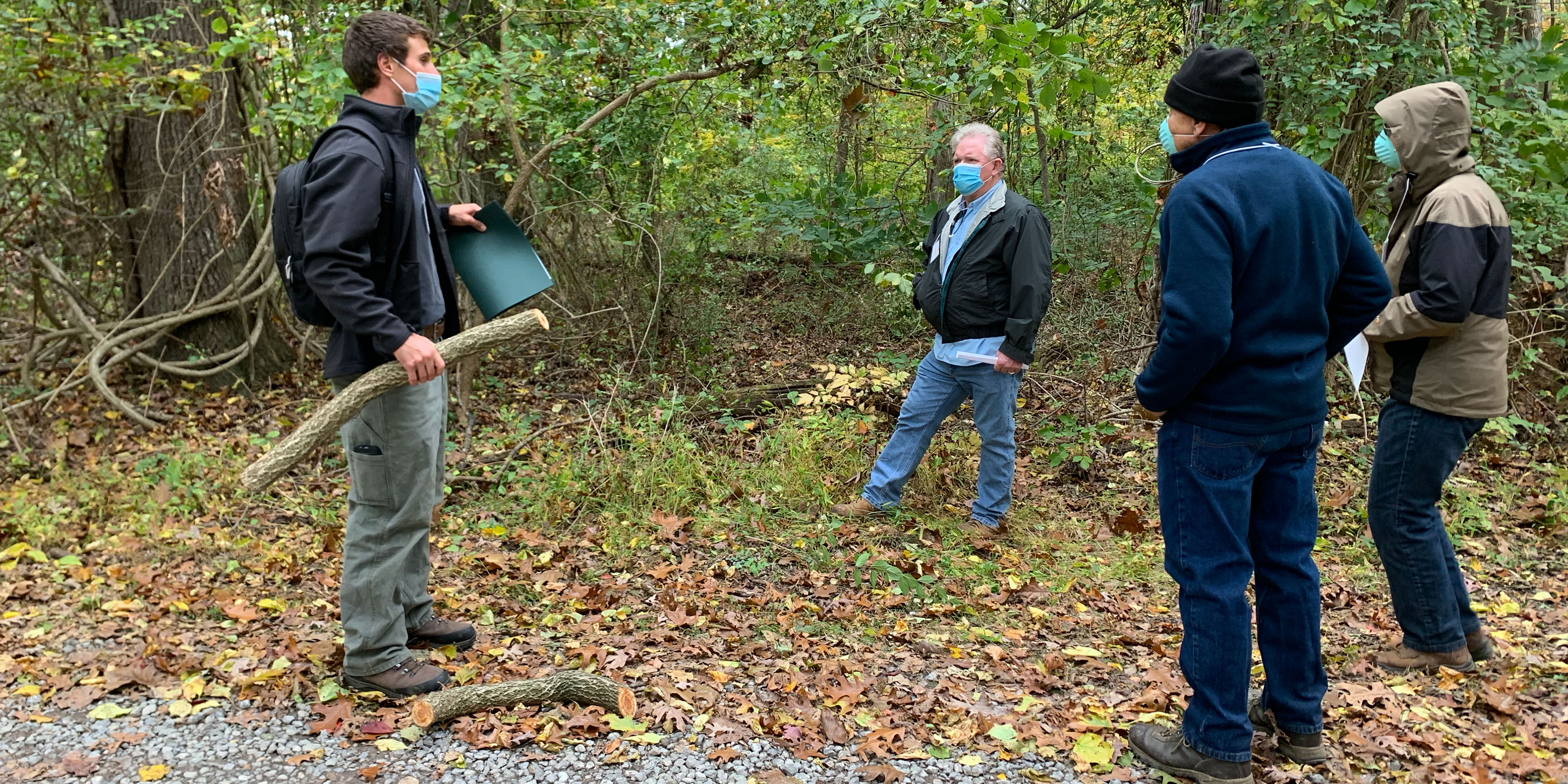 An instructor holding a three foot cut tree limb and folder standing with three participants wearing face coverings socially distanced outside between a road and forest.