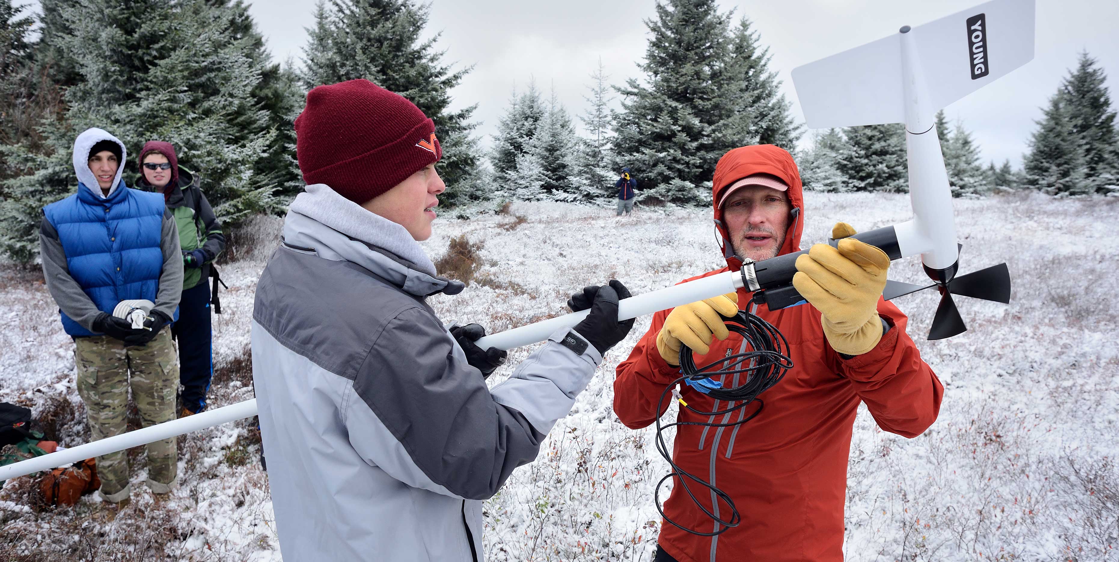 Two people in winter clothing hold a long piece of equipment. Two other people stand in the background watching them.