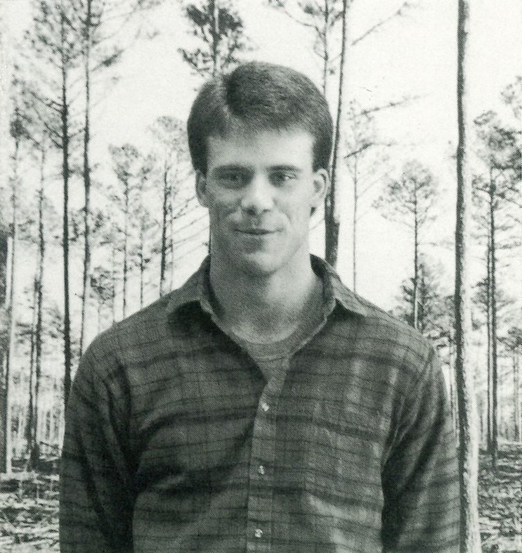 A black and white photo from 1989 of Easton Loving standing front of a backdrop of tall skinny pine trees.