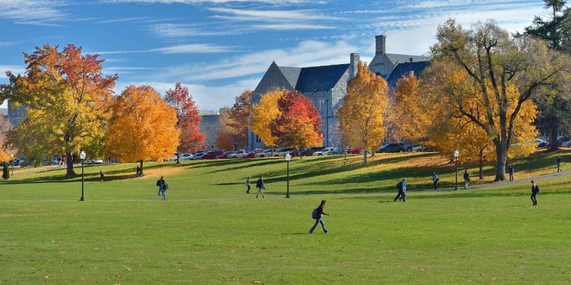 Several people walking across the Virginia Tech Drillfield with trees in fall color and campus buildings in the background.