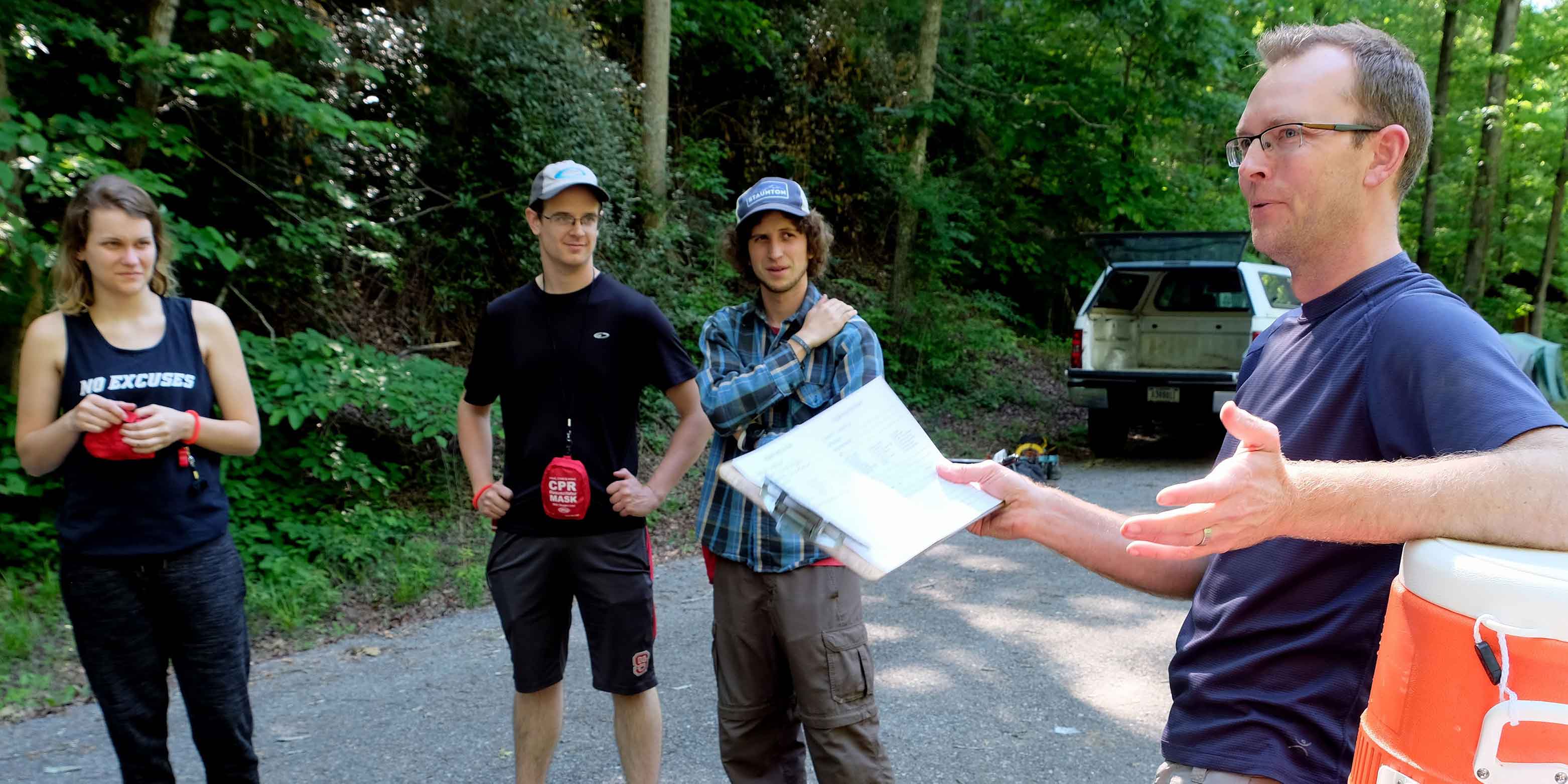 One woman and three men stand on a paved roadway in a forested area. One man holds a clipboard and addresses the other three.