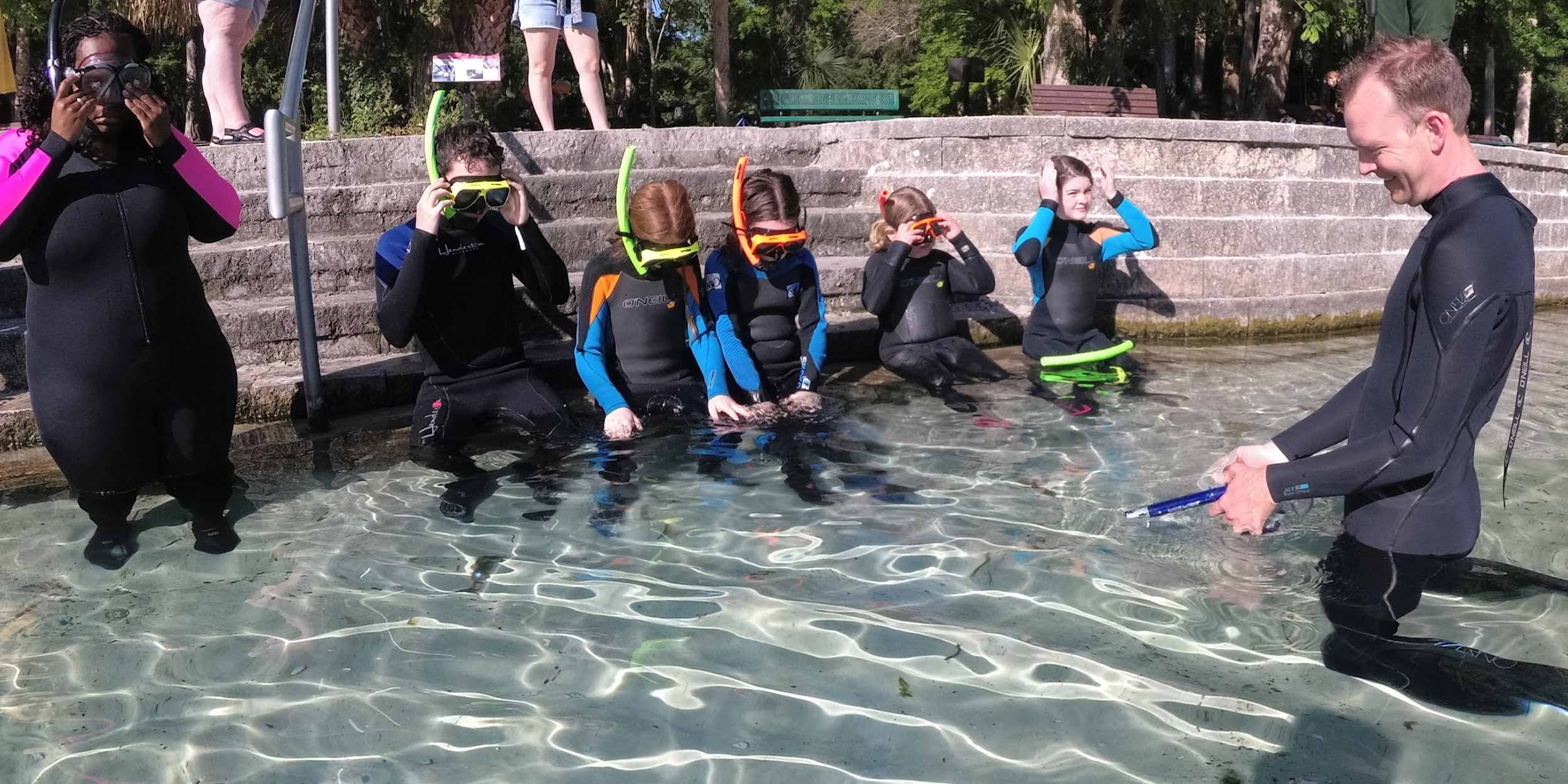 Several children in goggles, snorkels, and wetsuits sit on steps that extend into shallow, clear water. An adult, also in goggles, snorkel, and wetsuit, lays in the water faces them.