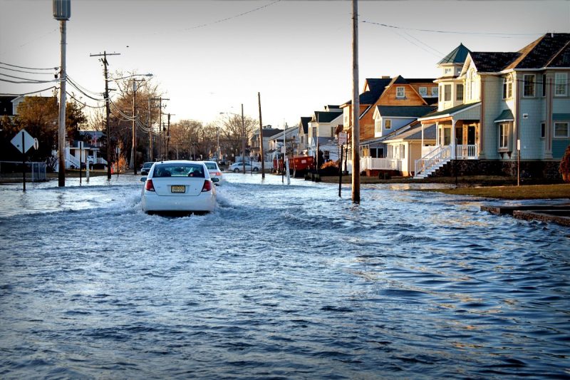 A white compact care traverses a flooded street in Hampton Roads, Virginia. The wake reaches is hitting telephone and electric poles that line the streets. The flooding is so high that the water reaches the porch steps of homes.