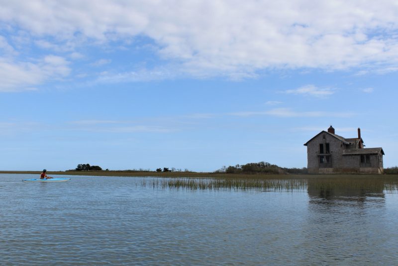 A man in a kayak floats near an abandoned house on the edge of a waterway.