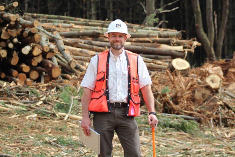 A man wearing a hard hat and an orange reflective vest and holding a clipboard stands in front of a large pile of cut logs