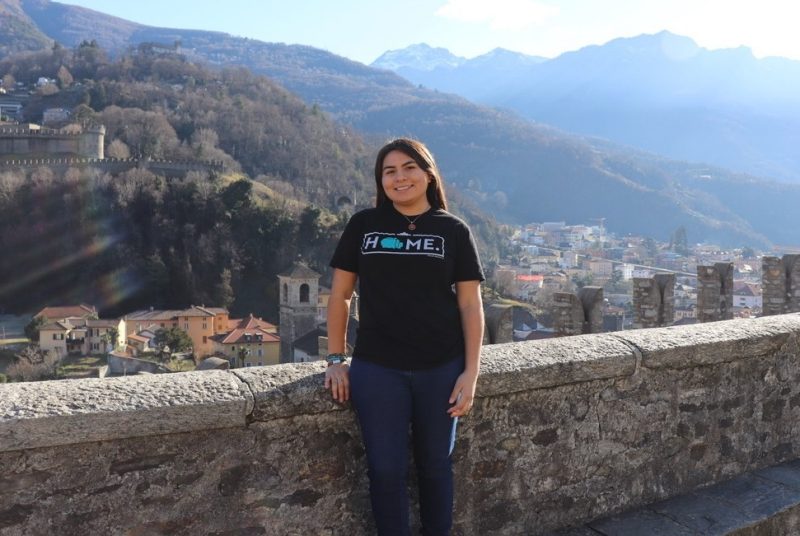 Nizhoni Tallas standing in front of a low rock wall with a village and a mountainous landscape behind her