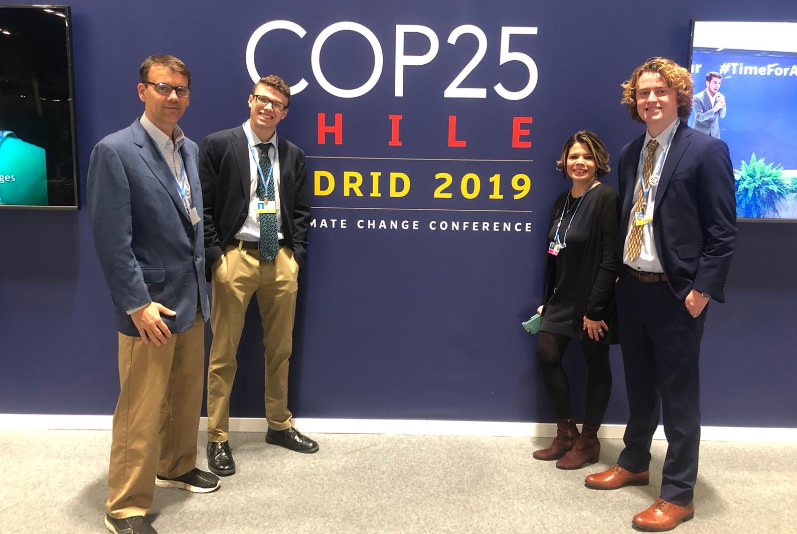 Three men and one woman standing in front of a large wall hanging for COP25 Chile Madrid 2019.