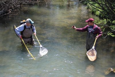 Two men wearing waders and holding fishing nets stand knee-deep in a stream. One facing the other while holding up a cellphone.