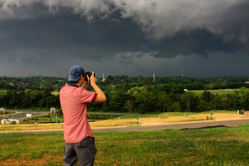 A man with his back to the camera holds a camera, looking towards dark clouds in the distance.