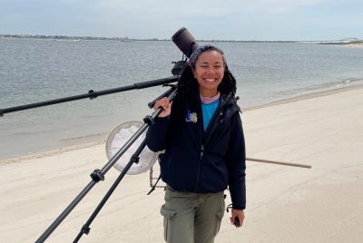 Woman carrying a spotting scope on a tripod over her shoulder while walking along a beach