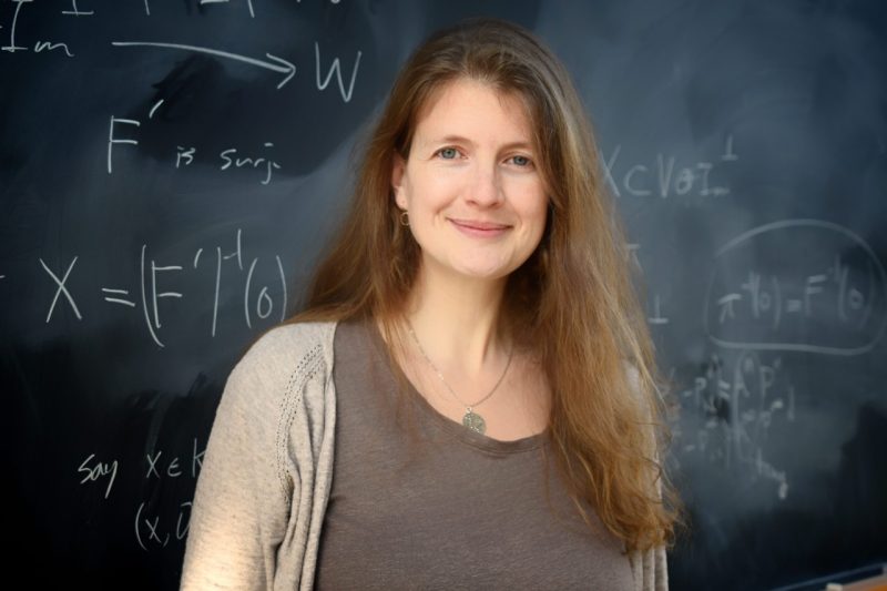 Lara Anderson, an associate professor of physics and affiliate professor of mathematics at Virginia Tech, stands in front of a chalkboard scrawled with math equations.
