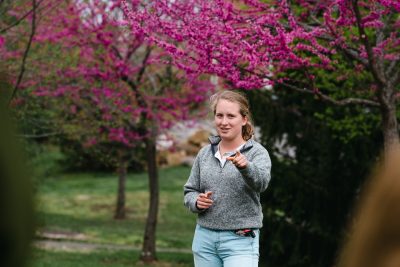 Grace Steger wearing a grey quarter zip jacket and light wash jeans points to the camera in front of flowering trees
